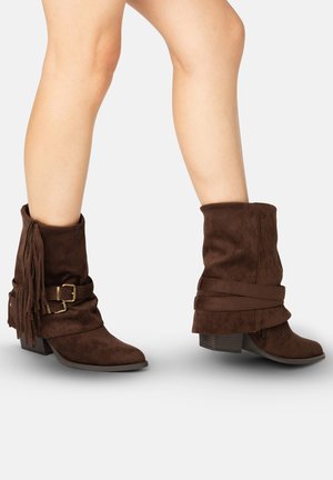 Bruine suède laarzen tot halverwege de kuit met lage hakken, decoratieve riemen, gespen en franjes op blote benen van een persoon.