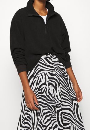 Person iført en sort quarter-zip trøje med op rullede ærmer og en sort-hvid zebra-print nederdel med højtaljet og knapper.