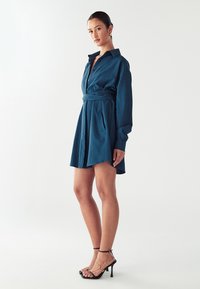 Temno zeleni srajčni obleki s ovratnikom, sprednjimi gumbi in zavezanem pasu. Ima dolge rokave in razširjen krilo. Ustrezajo ji črne sandale na peto.