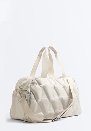 Kremowa pikowana torba podróżna z podwójnymi górnymi uchwytami oraz regulowanym paskiem na ramię na jednolitym białym tle.