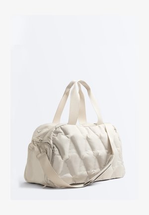 Cream-colored quilted duffel bag with dual top handles and an adjustable shoulder strap on a plain white background.