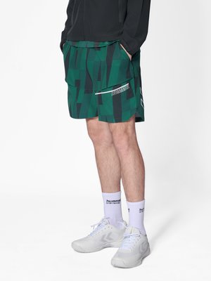 Mann trägt grüne und schwarz gemusterte Shorts mit "Hummel"-Logo, weiße Markensocken und weiße Sneakers, steht mit den Händen in den Taschen.