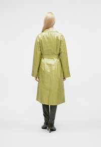 Lang, belteet trenchcoat i blank olivengrønt skinn, med bred krage og senkede skuldre, rett silhuett og vide ermer.