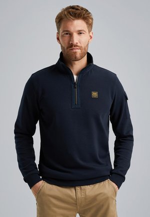 Felpa blu scuro con colletto a mezza zip, tessuto texturizzato, vestibilità slim e un piccolo emblema sul petto. Abbinata a pantaloni beige.