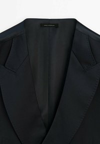 Černý přizpůsobený blazer s ostrými špičatými klopami, hladkým saténovým podšívkou a jemnými detailemi šití. Štítek uvádí "Vyrobeno v Portugalsku."