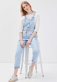 Salopette in denim azzurro chiaro con vita aderente, spalline regolabili, bottoni sul davanti e risvolti, abbinata a scarpe beige con lacci.