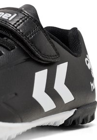 Chaussure de sport noire avec logo double flèche blanc, bande Velcro et semelle extérieure texturée conçue pour une utilisation en extérieur.