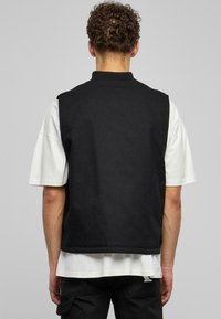 Melnas bezpaliktņa veste ar augstu apkakli, izgatavota no teksturētas auduma; valkāta virs baltas T-kreklu, ar brīvu piegriezumu un bez redzamām aizdarem.