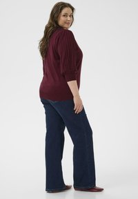 Pull en tricot côtelé bordeaux à manches trois-quarts, associé à un jean noir droit et des chaussures bordeaux, vu de côté.