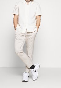 Camicia beige a maniche corte con bottoni, pantaloni beige a righe chiare e sneakers bianche con dettagli neri. Design casual e moderno.