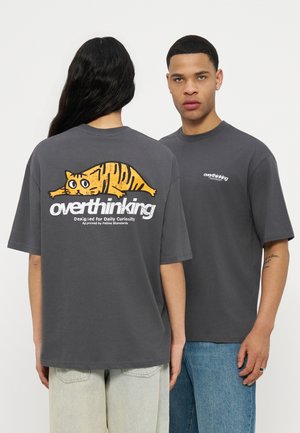 Twee personen dragen donkergrijze "overthinking" t-shirts; één toont de achterkant met kattengrafiek en tekst, de ander kijkt naar voren met een klein logo op de borst.