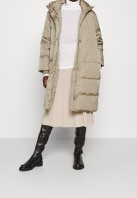 Beige, puffy lang jakke med hætte, vist åben over en hvid sweater og en plisseret beige nederdel, parret med sorte knæhøje støvler.