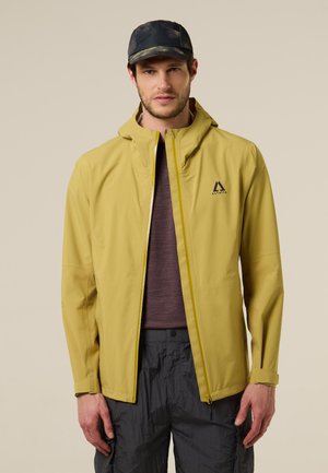 Hombre con una chaqueta amarilla con capucha sobre una camisa morada y pantalones negros, con una gorra negra, de pie contra un fondo beige.