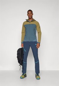 Lauko striukė alyvuogių žalios ir žydros spalvų, su užtrauktukais kišenėse ir prigludusiu kirpimu. Derinama su tamsiai mėlynomis kelnėmis ir tvirtais žygių batais.