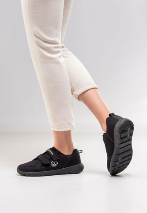 Osoba ubrana w beżowe spodnie i czarne buty typu slip-on z regulowanymi paskami, pokazująca wzór bieżnika podeszwy na jednej stopie.