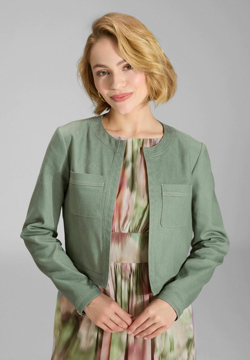 Jeune femme aux cheveux blonds courts et ondulés portant une veste courte vert sauge par-dessus une robe plissée aux couleurs pastel multicolores, posant les mains jointes.