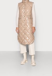 Beige quiltad puffer väst med glänsande yta, hög krage, två framfickor och tryckknappar, kombinerad med beige klumpiga stövlar och krämfärgade byxor.