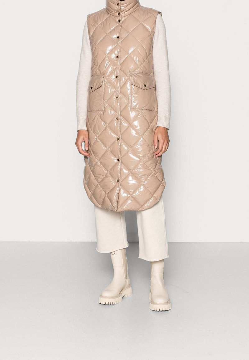 Beige quiltad puffer väst med glänsande yta, hög krage, två framfickor och tryckknappar, kombinerad med beige klumpiga stövlar och krämfärgade byxor.