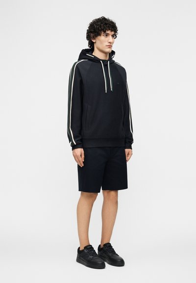 Νέος άνδρας που στέκεται, φοράει μαύρο φούτερ με κουκούλα και άσπρες ρίγες στα μανίκια, μαύρο σορτς και μαύρα αθλητικά παπούτσια, σε λευκό φόντο.