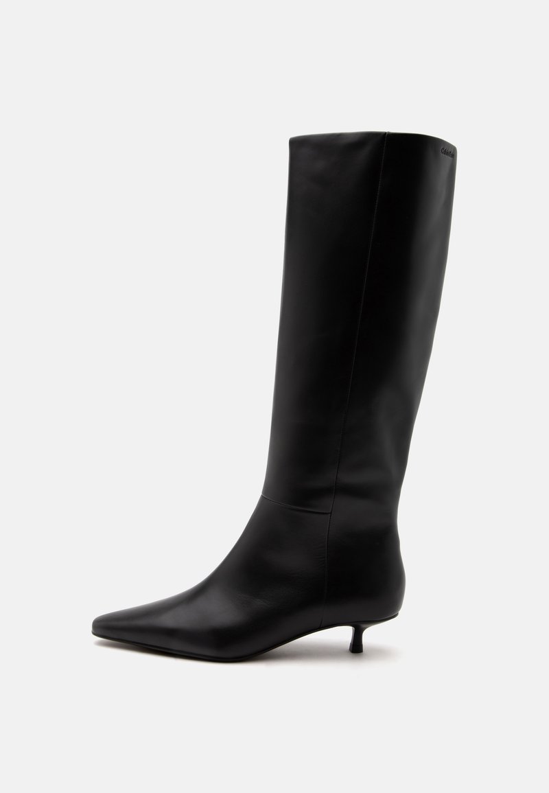 Botte noire en cuir jusqu'au genou avec bout pointu et petit talon kitten sur un fond clair uni.