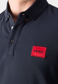 Gros plan d'un polo bleu marine avec un col côtelé, une patte de boutonnage et un écusson rouge portant la mention « HUGO » sur la poitrine.