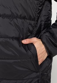 Czarna puchowa kurtka z gładkiego nylonu, z poziomymi szwami, kieszenią boczną i otworami na kciuki w rękawach. Teksturowana powierzchnia.