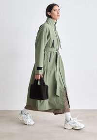 Cappotto lungo verde oliva con colletto alto, abbottonato in vita, dotato di cerniere nere e dettagli in contrasto. Abbinato a una borsa a tracolla scura e scarpe da ginnastica bianche.