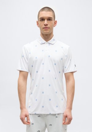 Ung mand med kort hår iført en hvid polo med små blå logoer og hvide shorts med sorte skospor på hvid baggrund.