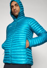 Teal-farbene, mit Kapuze versehene Steppjacke mit horizontalem Quiltmuster, Reißverschluss, zwei Seitentaschen und elastischen Bündchen, aus leichtem Material hergestellt.