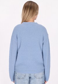 Pulover cosy de culoare albastră deschisă, cu guler ribbit, mâneci lungi și o textură moale, purtat peste o pereche de blugi deschiși la culoare, cu talie înaltă.