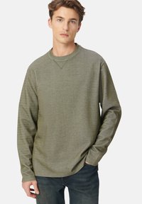 Junger Mann mit hellbraunen Haaren, der ein olivgrünes Langarmshirt und dunkle Jeans trägt, steht mit einer Hand in der Tasche.