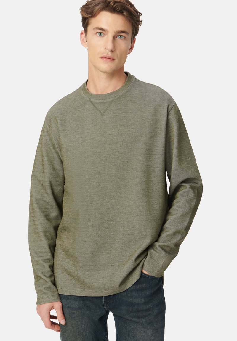 Junger Mann mit hellbraunen Haaren, der ein olivgrünes Langarmshirt und dunkle Jeans trägt, steht mit einer Hand in der Tasche.