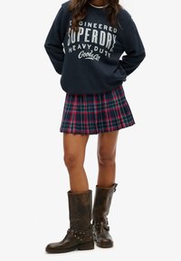 Sudadera de marina con logotipo blanco, falda de cuadros en azul marino, rojo y verde, y botas de cuero marrón con tachuelas plateadas y detalles de correa.