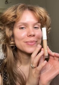 Un correcteur ILIA beige dans un tube transparent avec un capuchon blanc, tenu par une personne aux cheveux ondulés, de longueur épaule, sur un fond neutre.