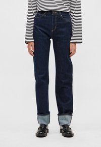 Tumedat sinist teksariide sirge säärega püksid, millel on heledates teksapükste värvitud ääred. Klassikalise viie taskuga disain kollaste õmblusdetailidega.