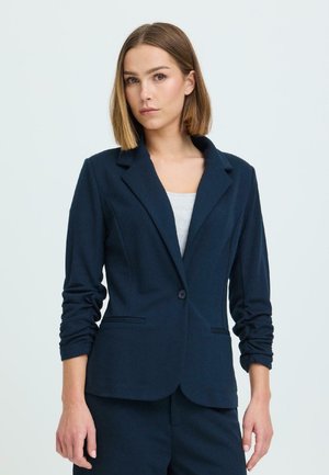 Naine, kellel on sirged õlgadeni juuksed, kannab tumesinist jopet ja helehallikat särke, taust on lihtne ja hele.