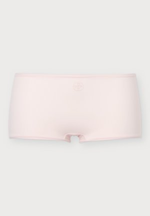 Mujer joven de pie descalza mirando hacia adelante, vistiendo un sujetador rosa claro con tirantes mínimos y ropa interior de talle alto a juego sobre un fondo liso.
