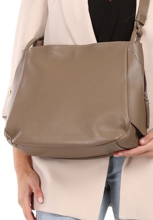 Sac à épaule en cuir taupe tenu par une personne portant une veste beige et un jean bleu, montrant une surface texturée et un détail de fermeture éclair.