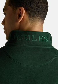 Zelená mikina s vysokým golierom. Golier má embosovaný nápis "JOULES." Tkanina vyzerá mäkká a textúrovaná, so čistým finišom.