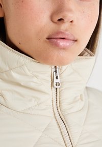 Lys beige pufferjakke med høj krave, der har en synlig lynlås og quiltet mønster for tekstur. Nærbillede af krave og halsudskæring.