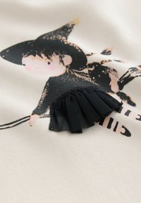 Tissu blanc avec une illustration fantaisiste d'une sorcière ; détail de tissu noir plissé en bas ; texture douce et design ludique.