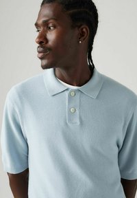 Vaaleansininen neulottu polo-paita lyhyillä hihoilla, jossa on kaksi vaaleaa nappia ja teksturoitu kangas, esiteltynä mallilla.