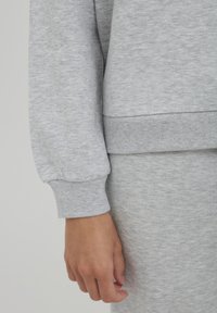 Tissu de sweat-shirt gris avec une texture douce ; doté de poignets et d'un ourlet côtelés, et d'une coupe décontractée. Main visible, avec une peau lisse.