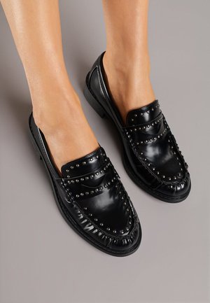 Ballerines vernies noires avec des clous argentés portées pieds nus sur un fond gris, mettant en valeur les détails et l'ajustement des chaussures.