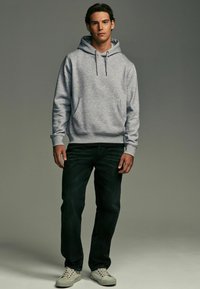 Gray hooded sweatshirt with a kangaroo pocket, paired with dark jeans and light gray sneakers. The fabric appears soft and textured.
