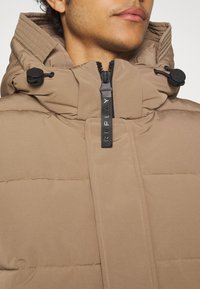 Giacca imbottita marrone con colletto alto, chiusura con zip e dettagli regolabili con cordini al collo. Presenta cuciture orizzontali e una texture morbida.
