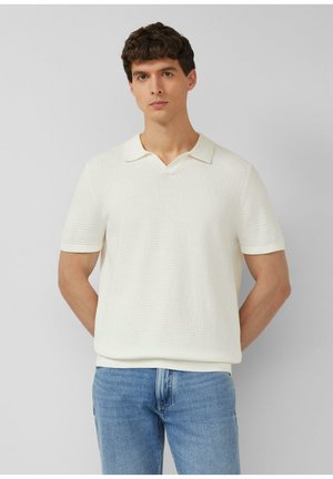 Młody mężczyzna stojący z rękami za plecami, ubrany w białą teksturowaną koszulkę polo z krótkim rękawem i niebieskie dżinsy, neutralne tło.