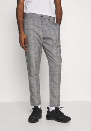 Mann trägt graue karierte Slim-Fit-Hosen, weiße Socken, schwarze Sportschuhe und ein weißes T-Shirt, steht vor einem weißen Hintergrund.