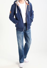 Sudadera con cremallera de color marino con un capucha forrada de felpa y cordones blancos, combinada con jeans de color azul claro y zapatillas blancas.