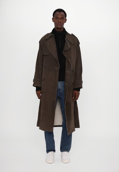 Brauner, langer, doppelt geknöpfter Trenchcoat mit breiten Revers, Seitentaschen und umgeschlagenen Ärmeln, getragen über einem schwarzen Oberteil und Jeans.